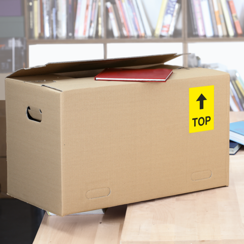 Una caja de cartón con un cuaderno rojo en la parte superior, etiquetada con un letrero amarillo que dice 'TOP', está sobre una mesa en una biblioteca.